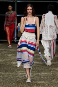 Model on a runway wearing a blue, red, white striped tank top and matching striped skirt, with white sneakers and a colorful knit bag.