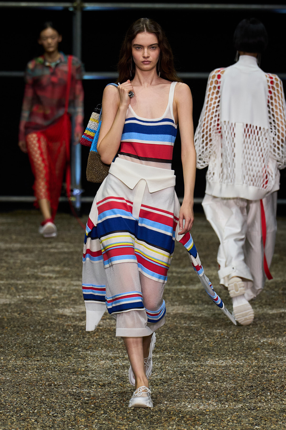 Model on a runway wearing a blue, red, white striped tank top and matching striped skirt, with white sneakers and a colorful knit bag.