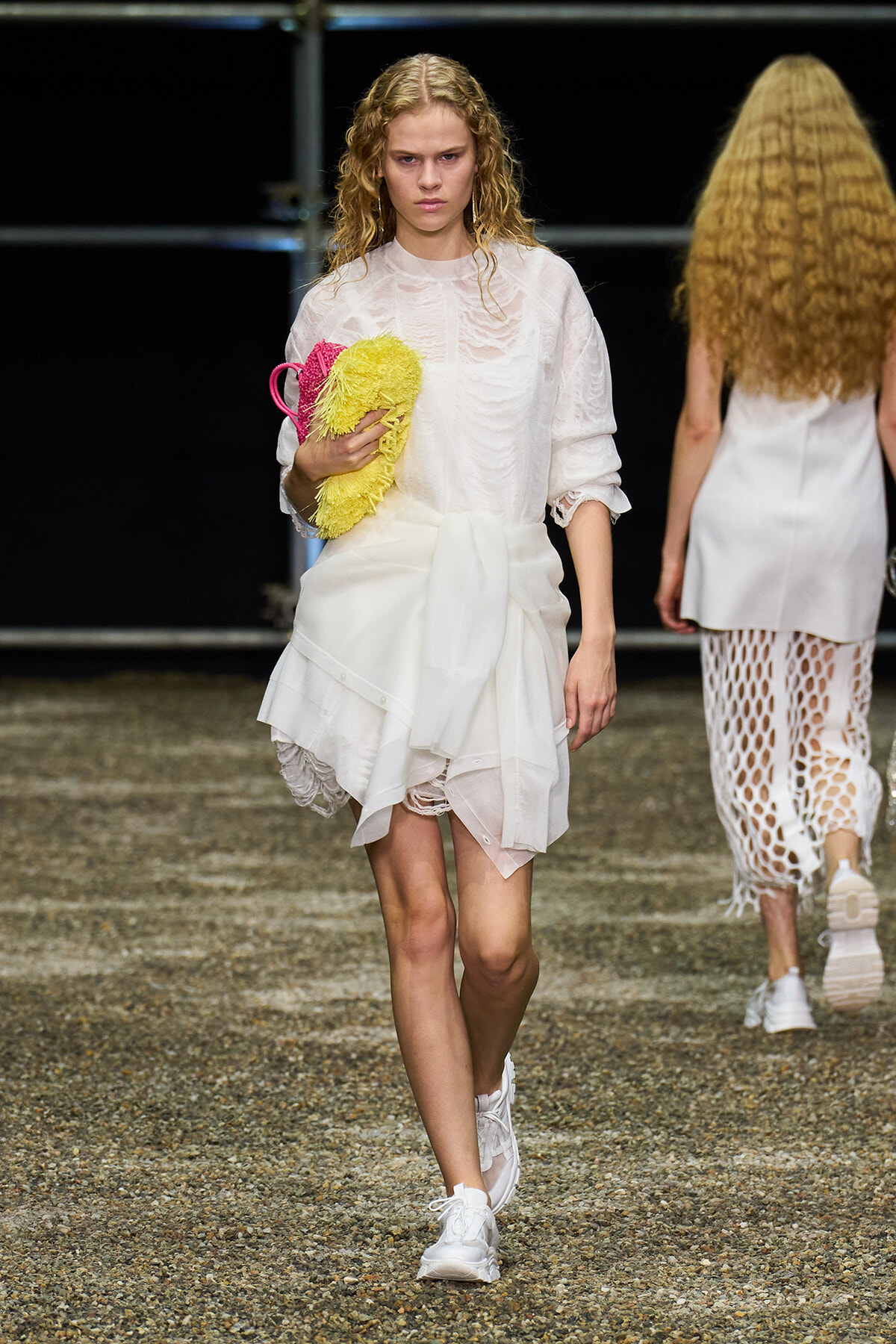 Model walking a runway in a white textured dress, holding a bright yellow fluffy clutch-like accessory.