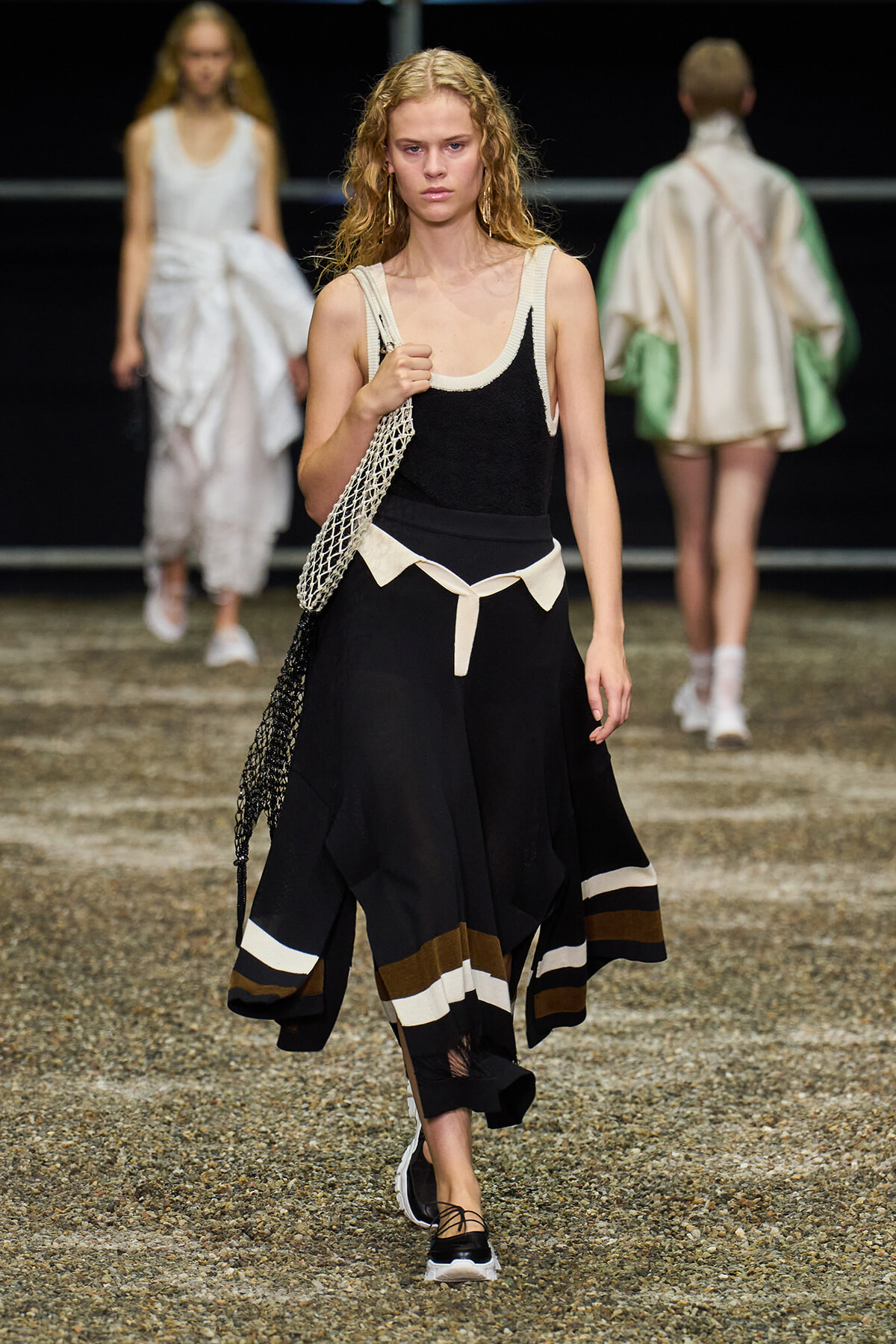 Model walking a runway in a black sleeveless dress with white trim, a cream belt, and a mesh tote bag over her shoulder.