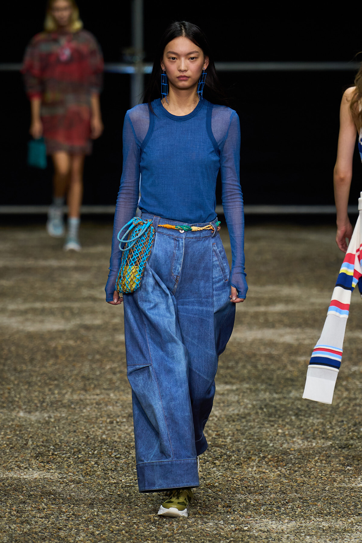Model walking the runway in a blue sheer long-sleeve top and wide-leg denim pants, with a blue net bag at the hip and bold blue earrings.