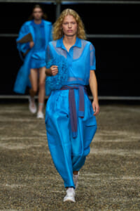 Model walking a runway in a bright blue outfit, holding a blue fluffy circular accessory.