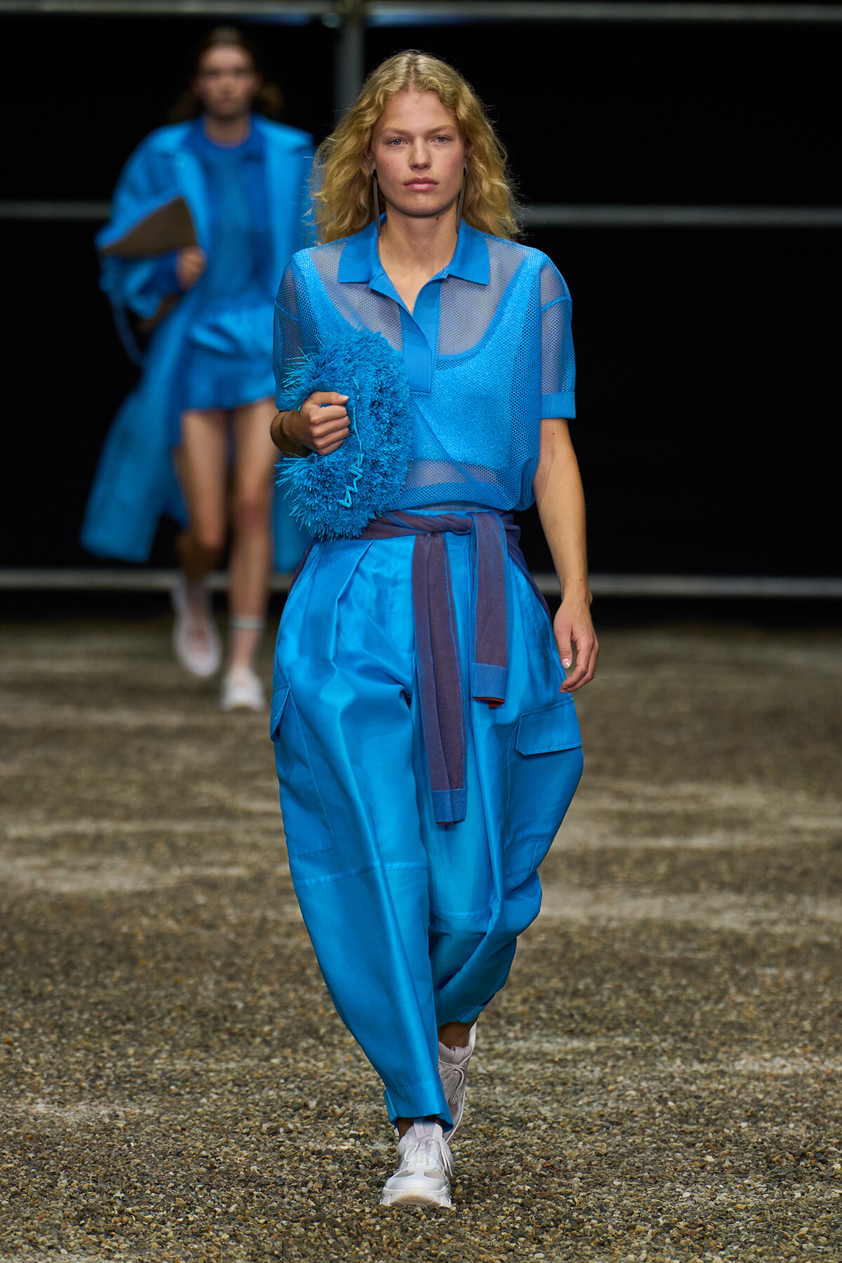 Model walking a runway in a bright blue outfit, holding a blue fluffy circular accessory.