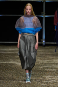 Model on a runway wearing a sheer blue color-block top over a cobalt layer, paired with a loose gray midi skirt, turquoise belt, and teal sneakers with socks.