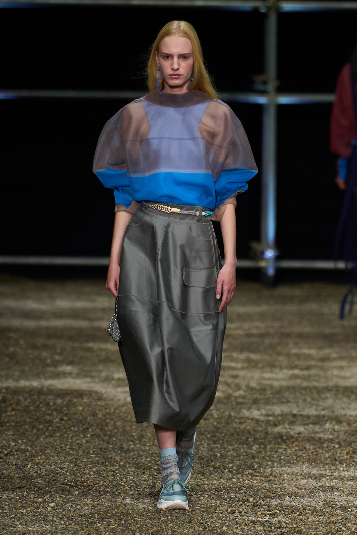 Model on a runway wearing a sheer blue color-block top over a cobalt layer, paired with a loose gray midi skirt, turquoise belt, and teal sneakers with socks.