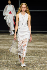 Runway model in a white sleeveless dress with open-weave mesh leggings, carrying a silver mesh tote bag.
