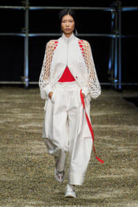 Model walking the runway in a white mesh zippered cape over a red top and oversized white cargo pants with a red stripe detail
