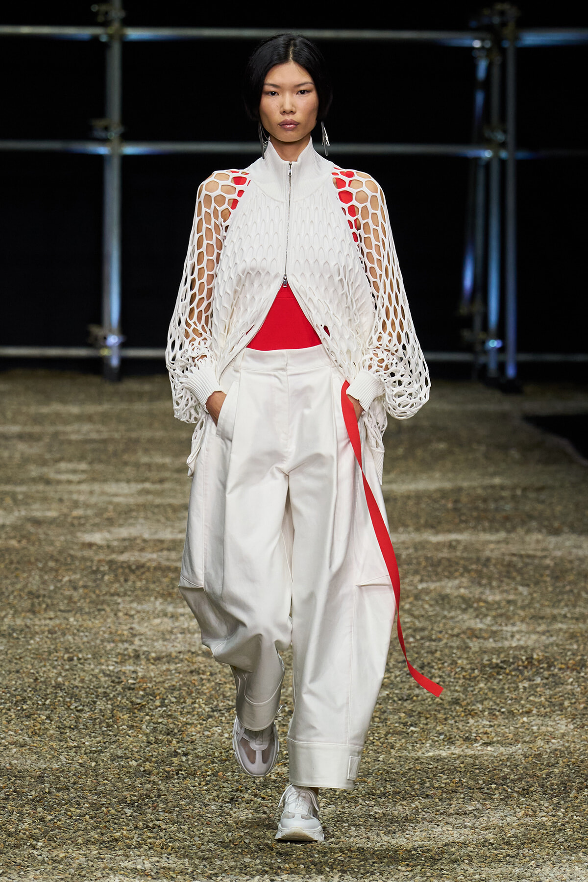 Model walking the runway in a white mesh zippered cape over a red top and oversized white cargo pants with a red stripe detail