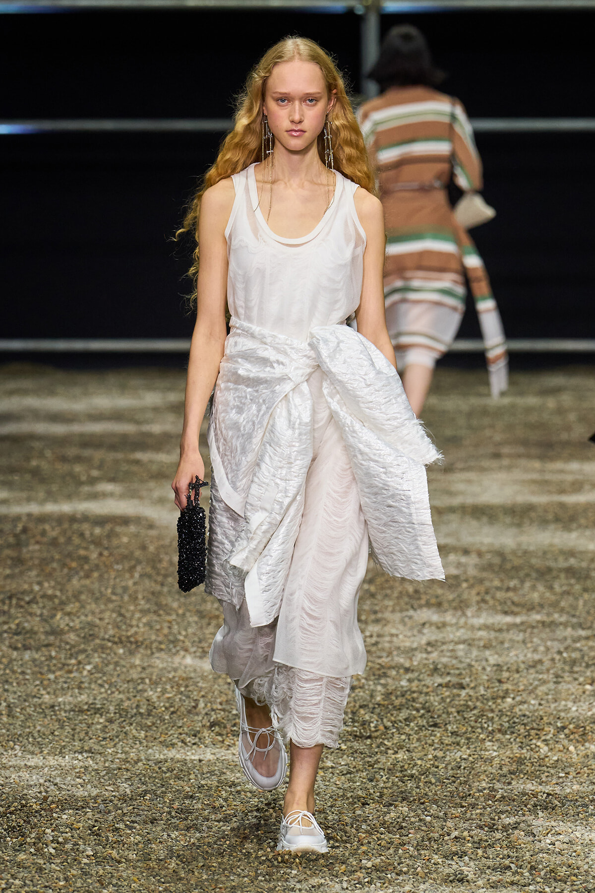 Model walks a runway in a white layered dress, white sneakers, and a small black beaded clutch with long wavy hair.