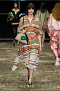 Model walking the runway in a striped tan, green, and white dress with belt, carrying a woven bag with green accent and black crossbody strap.