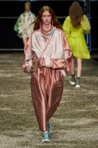 Model walks a runway in a pink satin oversized tracksuit with a beaded crossbody bag and light teal sneakers, spotlight on fashion show.