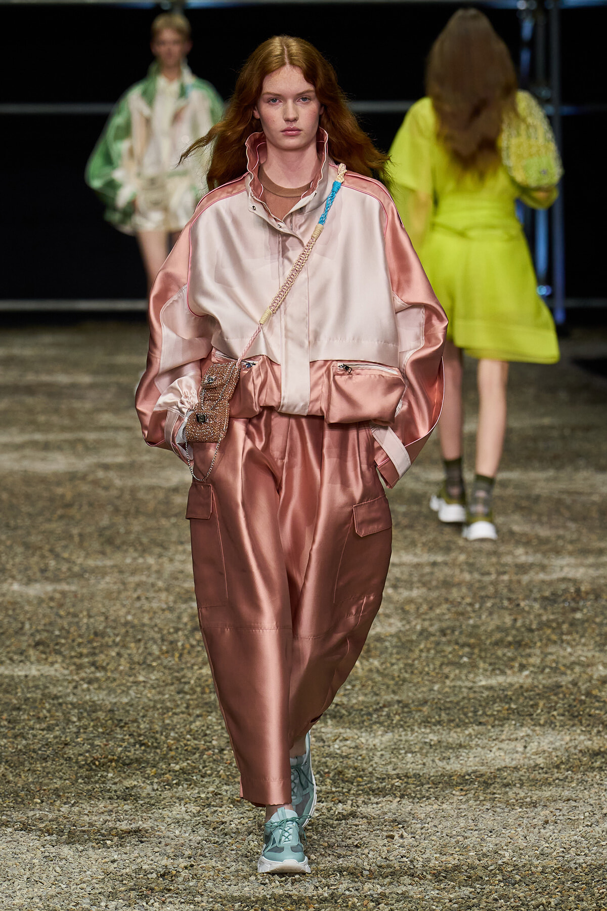 Model walks a runway in a pink satin oversized tracksuit with a beaded crossbody bag and light teal sneakers, spotlight on fashion show.