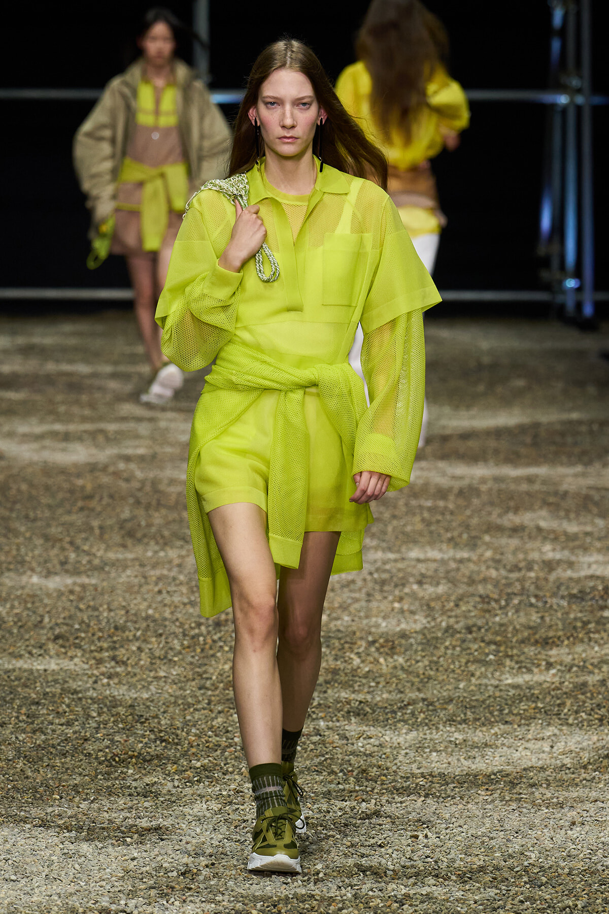 Model walks a fashion runway in a neon yellow sheer shirt-dress with a tied waist, holding a metallic rope accessory over her shoulder.