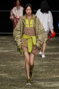 Model walking on a runway in a beige oversized jacket, lime-green layers, and tan dress, carrying a neon green accessory.