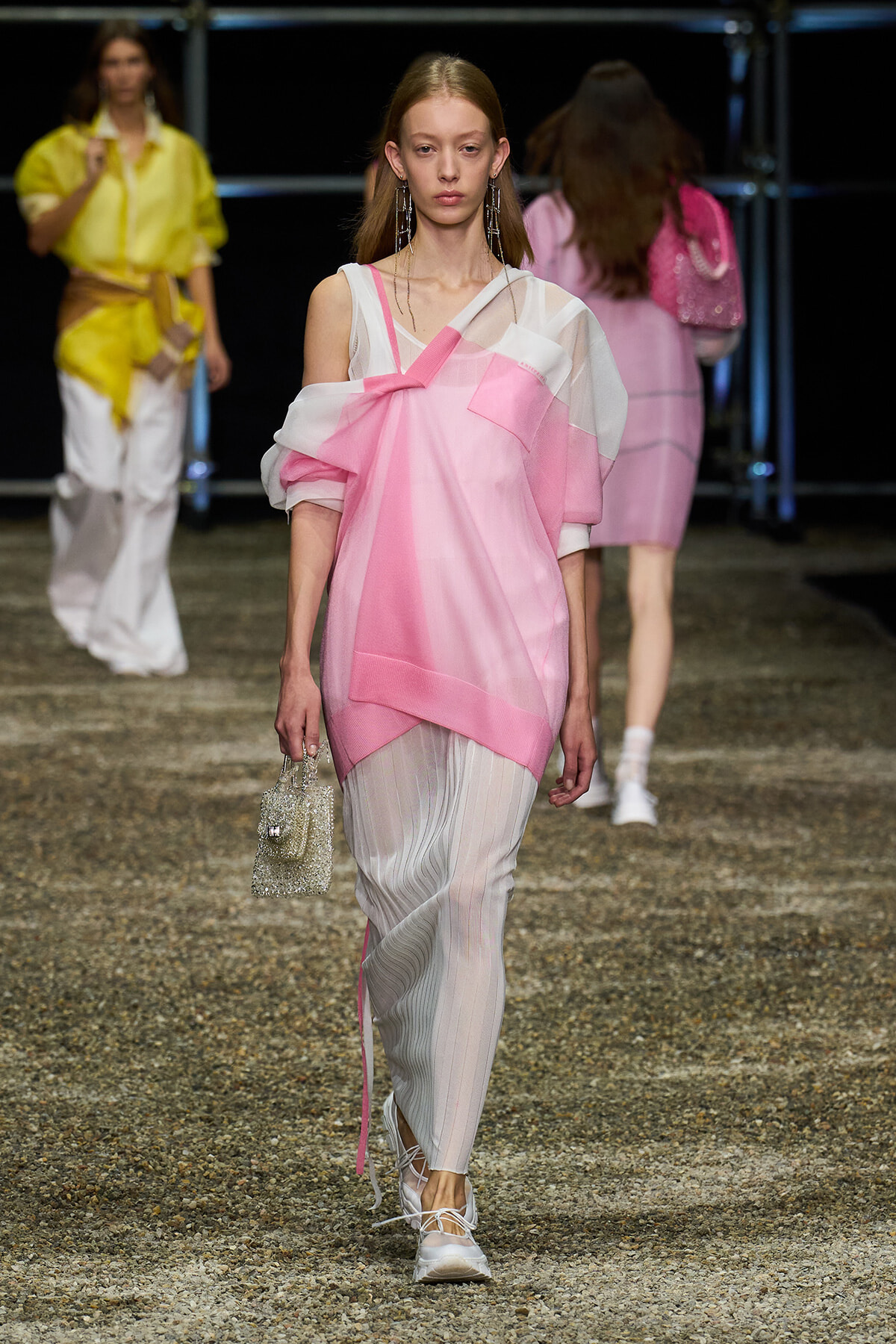 Female model walking a fashion show runway in a pink and white layered top over sheer striped pants, carrying a small glittery handbag and wearing long earrings.
