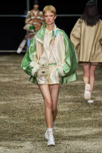 Model walks a fashion runway in a mint-green and cream satin jacket over a cream shirt and shorts, with a small woven crossbody bag and white sneakers.