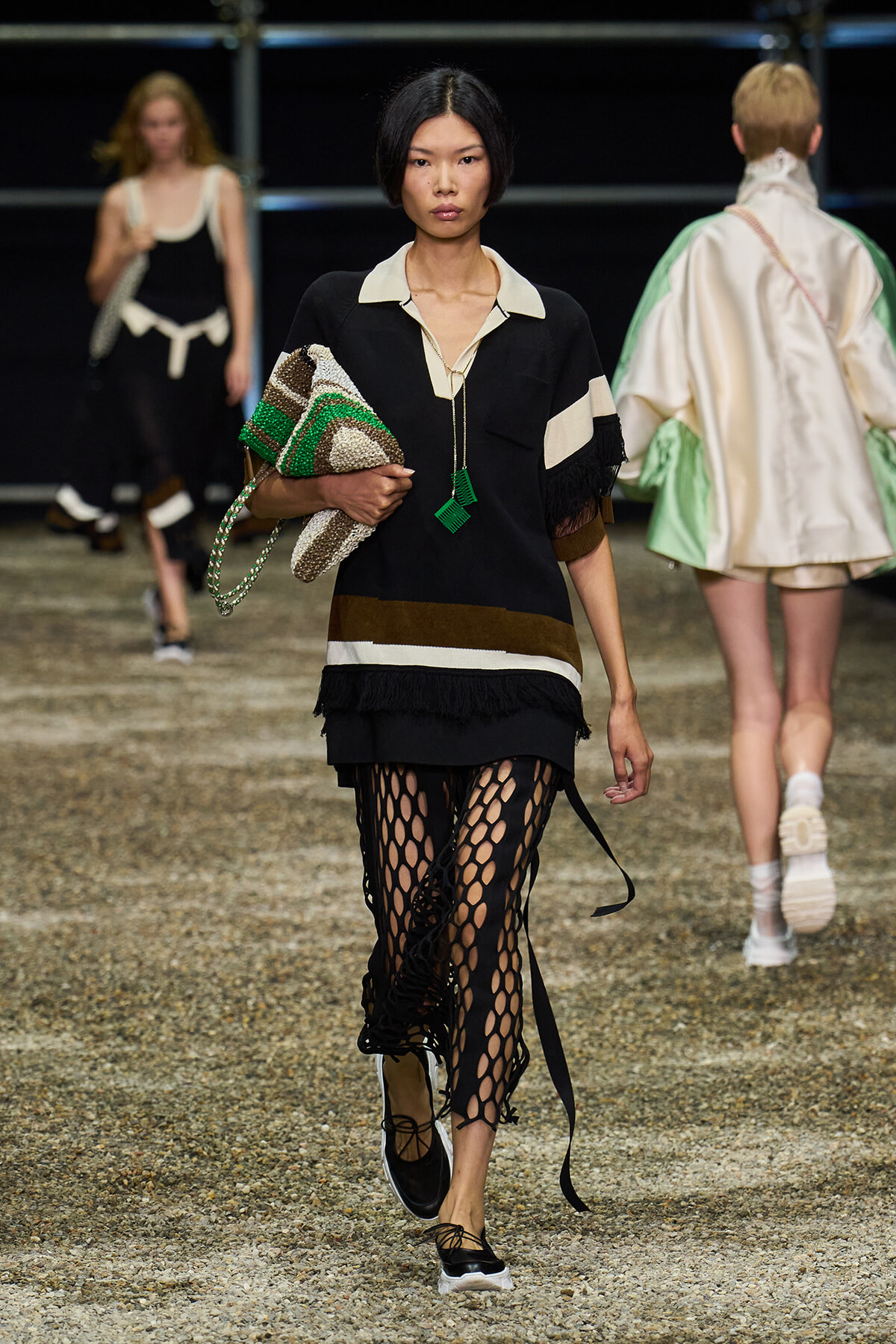 Model walking a runway in a black layered tunic with white collar, holding a textured woven bag, wearing fishnet leggings and black sneakers.