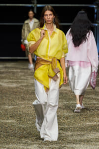 Model walks a fashion runway in a bright yellow sheer blouse cinched with a fabric belt over wide white pants, looking straight ahead.