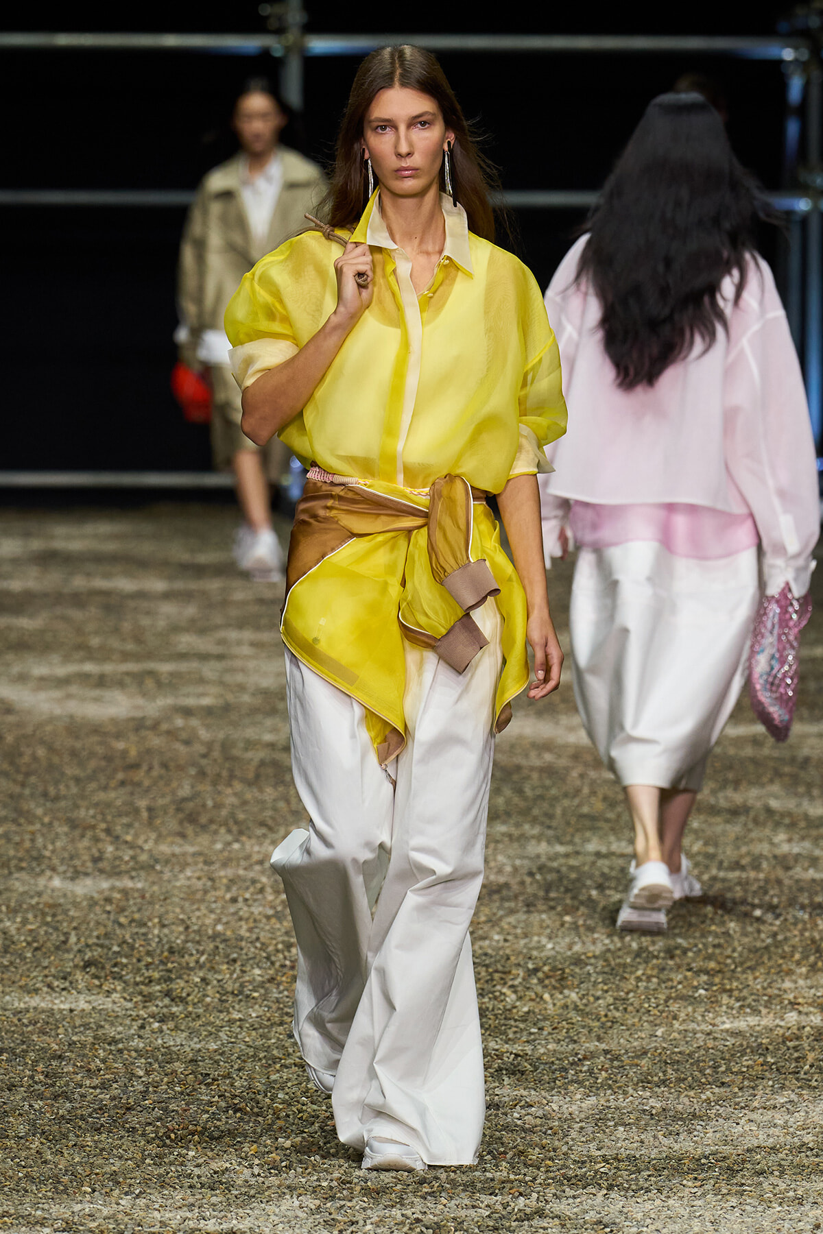 Model walks a fashion runway in a bright yellow sheer blouse cinched with a fabric belt over wide white pants, looking straight ahead.
