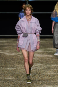 Model walks a runway in a lavender oversized coat with a belt, matching mini skirt and a silver belt bag