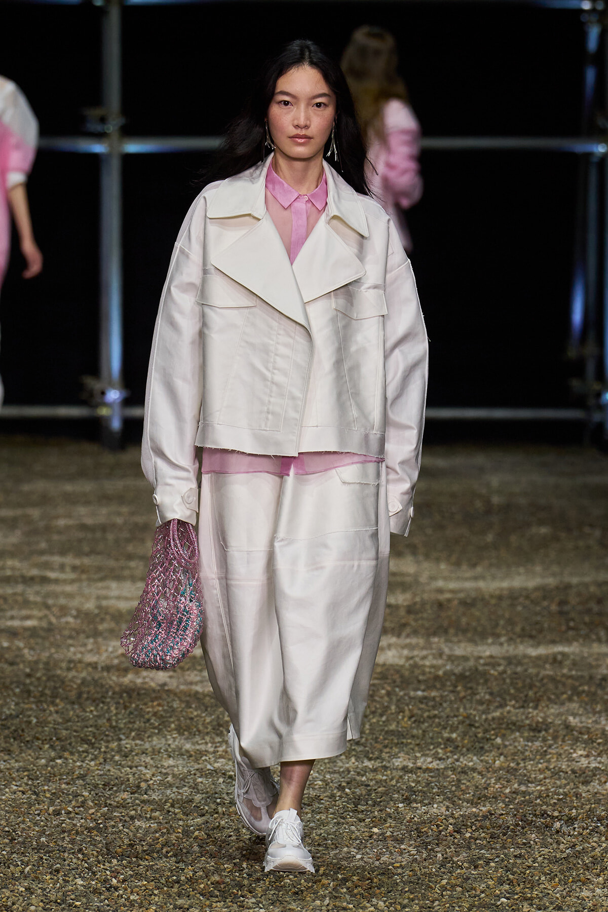 Model walks a runway in an oversized white jacket over a pink shirt, paired with white wide-leg pants and a pink mesh bag.