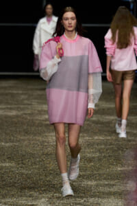 Female model walking a pink color-blocked tunic with mesh sleeves on a gravel runway, carrying a pink bag over the shoulder.