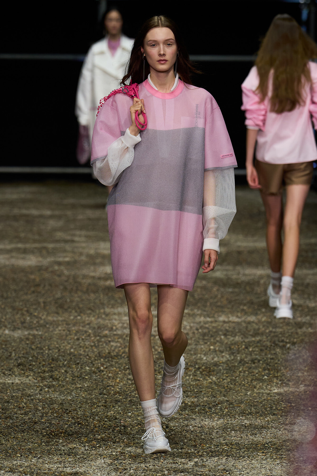 Female model walking a pink color-blocked tunic with mesh sleeves on a gravel runway, carrying a pink bag over the shoulder.