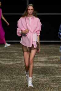 Model walking a fashion runway in a loose pink button-down shirt, brown shorts, white sneakers, and a silver crossbody bag.
