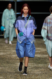 Model walking a runway in a blue-purple color-block windbreaker, layered denim shorts, black socks, sneakers, and a turquoise crocheted crossbody bag.