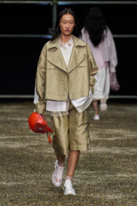 Model walking a runway in a beige oversized jacket, white shirt, knee-length shorts, and white sneakers, carrying a red mini handbag.