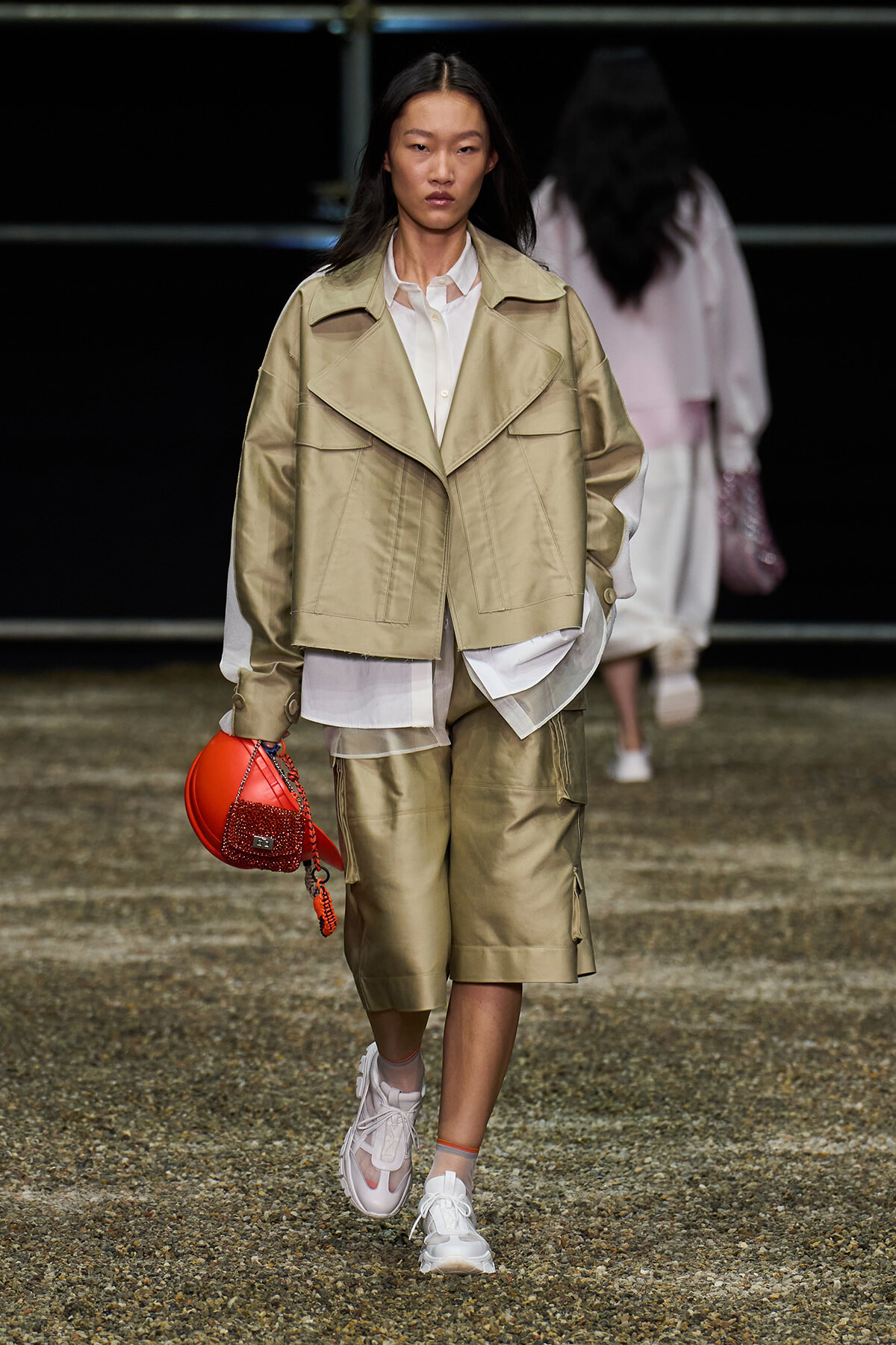 Model walking a runway in a beige oversized jacket, white shirt, knee-length shorts, and white sneakers, carrying a red mini handbag.