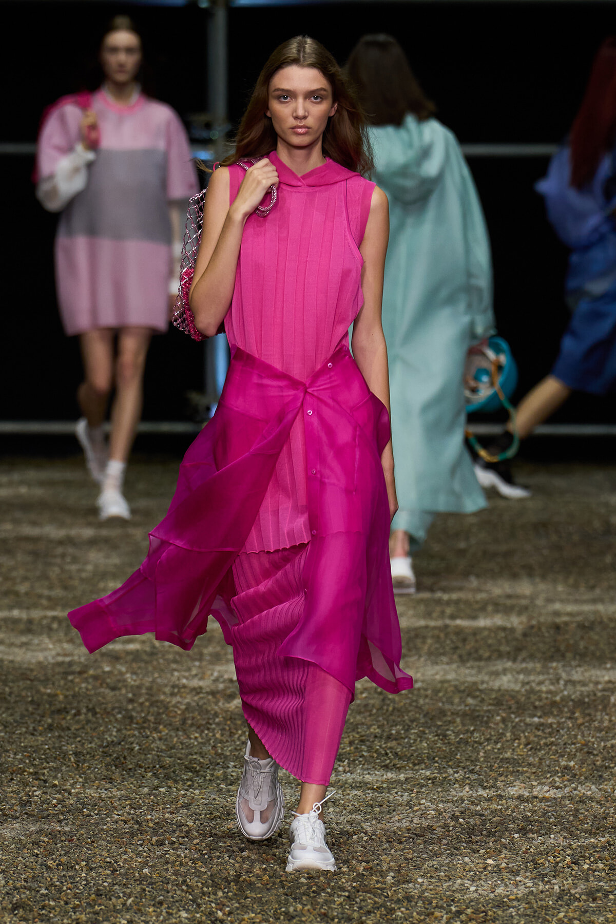 Model walking a runway in a vivid pink sleeveless dress with a layered sheer skirt and white sneakers, carrying a pink chain-strap bag.