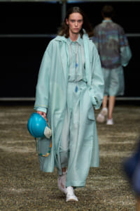 Model walks the runway in a mint trench coat and matching wide-leg pants, white sneakers, and a turquoise hard hat in hand.