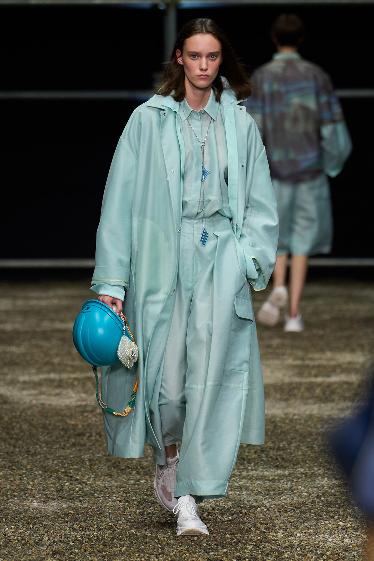 Model walks the runway in a mint trench coat and matching wide-leg pants, white sneakers, and a turquoise hard hat in hand.