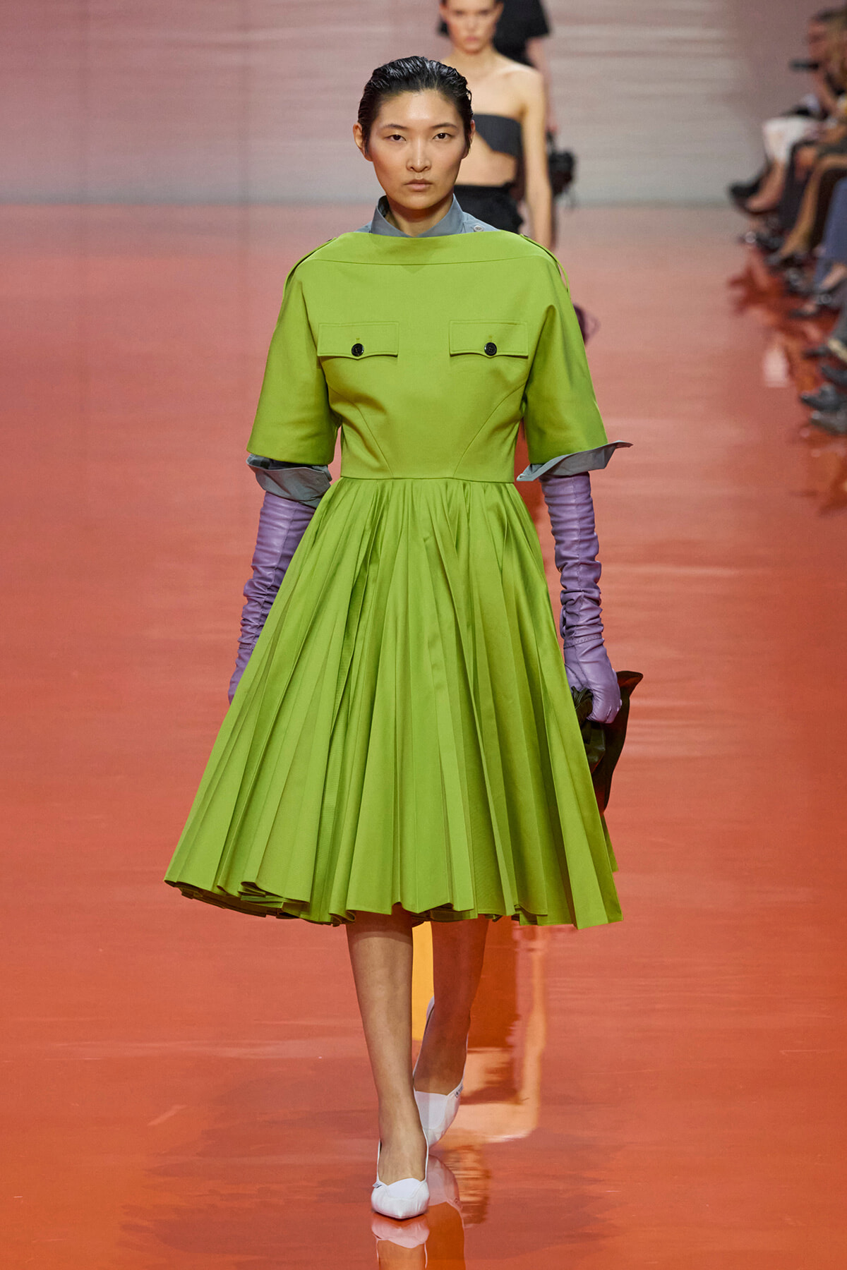 Model walks down a glossy orange runway wearing a bright lime green pleated dress with short sleeves, lavender gloves, and white heels.