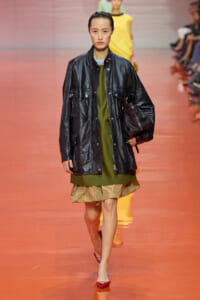 Model on a runway wearing an oversized black leather jacket over an olive-green dress with a beige ruffled hem, carrying a black bag and wearing red heels.