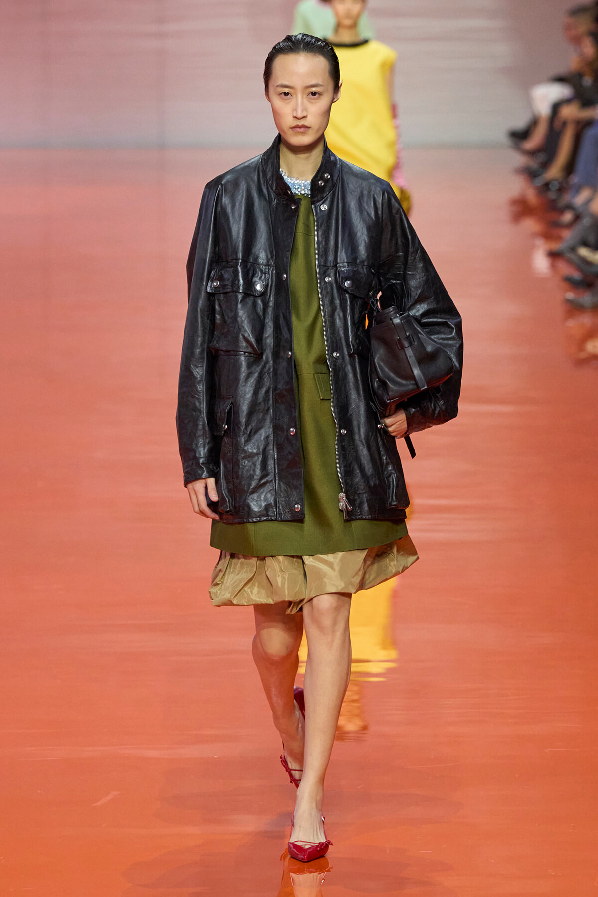 Model on a runway wearing an oversized black leather jacket over an olive-green dress with a beige ruffled hem, carrying a black bag and wearing red heels.