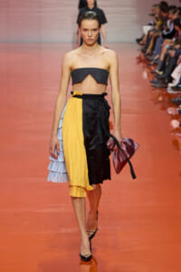 Model walking a fashion runway wearing a color-blocked skirt with a yellow and black wrap, strapless gray top, and black heels.