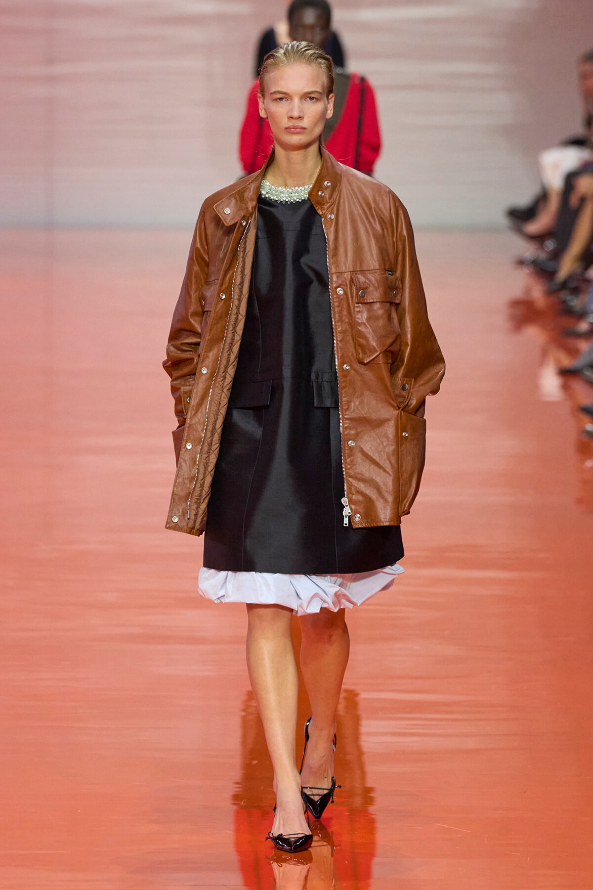 Model walking a runway in a brown leather utility jacket over a black dress with a white ruffled hem, on a glossy pink runway.