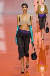 Model walks a pink-orange runway in a beige bralette, teal arm-warmers, sheer black pants with a purple waistband, and a small black handbag.