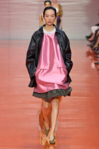 Model walking a fashion runway in a pink satin dress with a black leather jacket and white pearl neckline, center stage.