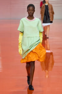 Model walking a runway in a mint-green dress with a pink beaded collar, long yellow gloves, and an orange ruched hem.