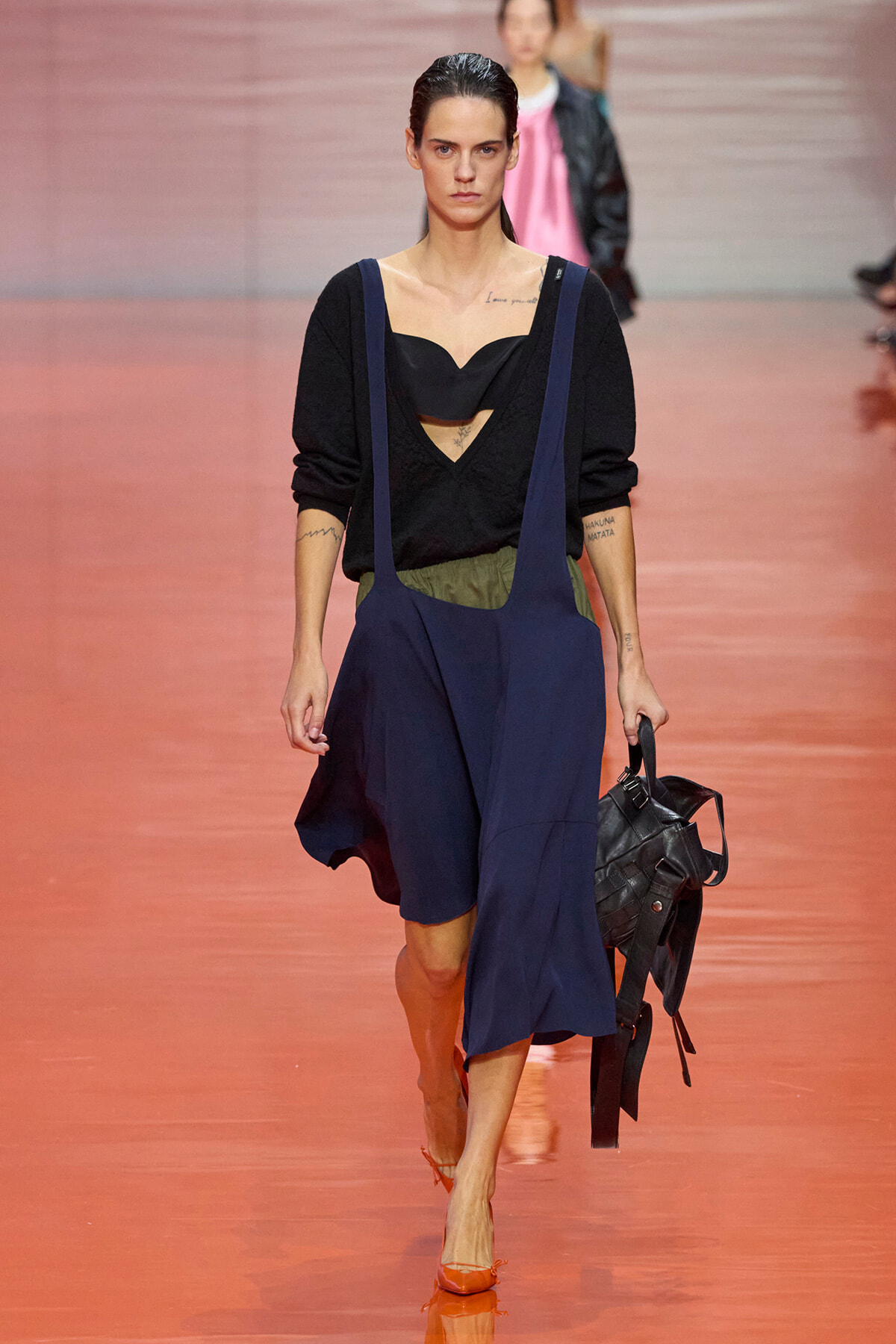 Model walking a fashion runway in a dark blue suspender skirt over a black top, orange heels, and carrying a black handbag.