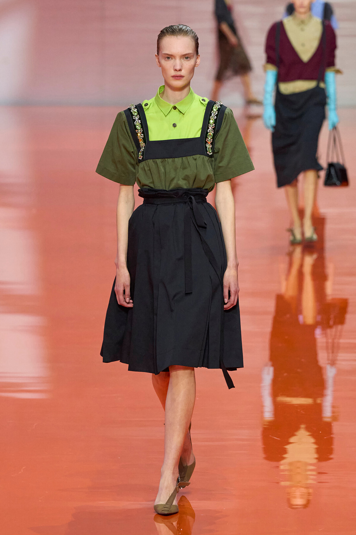 Model walking the runway in a lime green collared top with olive green embellished straps and a black wrap skirt.