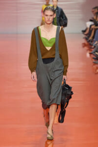 Model walking a runway in a lime green top under a brown V-neck sweater and gray suspender pants, carrying a black bag on her side.