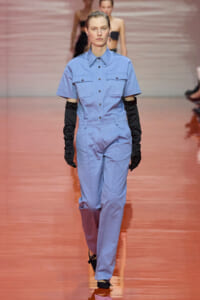 Model walking a fashion runway in a light blue short-sleeve utility jumpsuit, paired with long black gloves and black heels.
