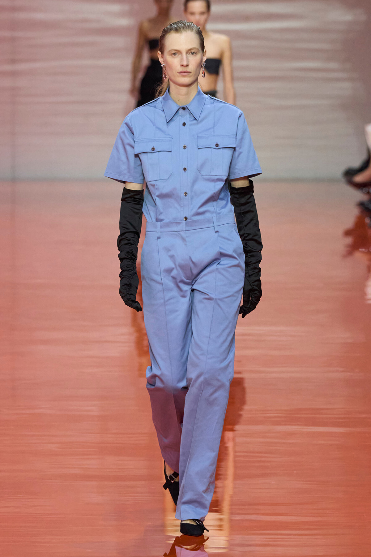 Model walking a fashion runway in a light blue short-sleeve utility jumpsuit, paired with long black gloves and black heels.