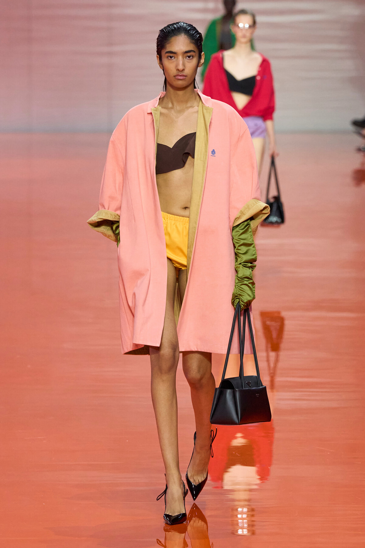 Model walks the runway in a pink oversized coat with olive green sleeves, yellow shorts, and a brown bandeau top, carrying a black handbag and wearing black heels.
