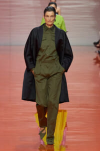 Model walks the runway wearing an olive-green utility shirt and trousers with a black cape draped over the shoulders.