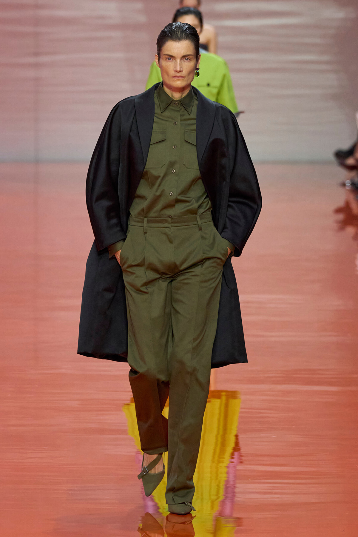Model walks the runway wearing an olive-green utility shirt and trousers with a black cape draped over the shoulders.